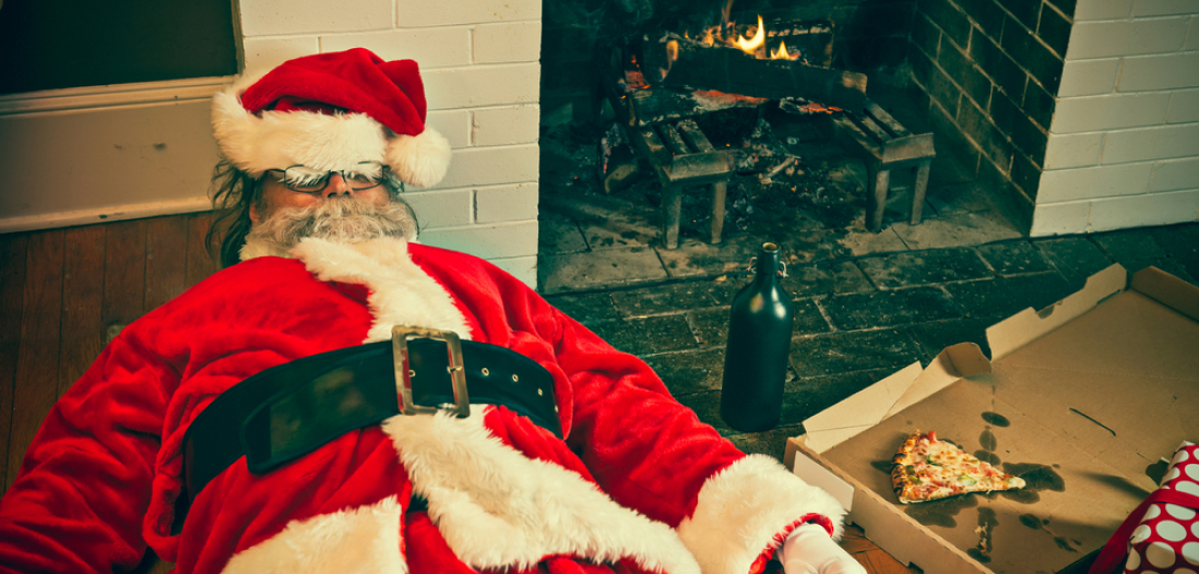 Santa Clause sleeping by the fireplace with an empty pizza box and empty bottle beside him